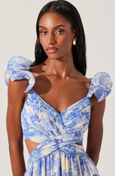 A woman is wearing a light blue and white patterned dress with ruffled shoulder straps, a sweetheart neckline, and cutouts at the waist. The dress has a pleated bodice and a flowing skirt with a matching pattern.